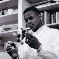 A photo angled up of a dark skinned young man wearing a lap coat and holding a molecule structure with both of his hands.