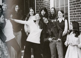 A group of fair skinned female students stand on the steps of a brick building, laughing and smiling together.