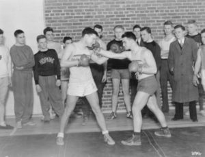 Group photograph of two Johns Hopkins University students boxing. A small crowd of other students stand around and watch them.