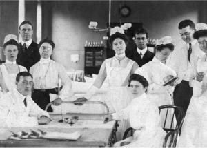 A group of white women nurses in early 20th century nurse uniforms stand around a table where a man where a doctor's jacket and a women wearing a similar nurse uniform sit down. There are two other men who stand behind the group of women wearing dark suits.