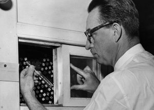 A white man wearing glasses examines a test tube pulled from a full of individual test tubes.