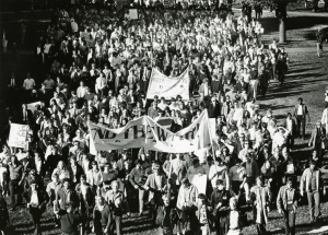 An overhead shot of a large crowd of people walking on university grounds. Demonstrators hod up signs and banners. The largest banner at the front has the words End the War, partially obscured.