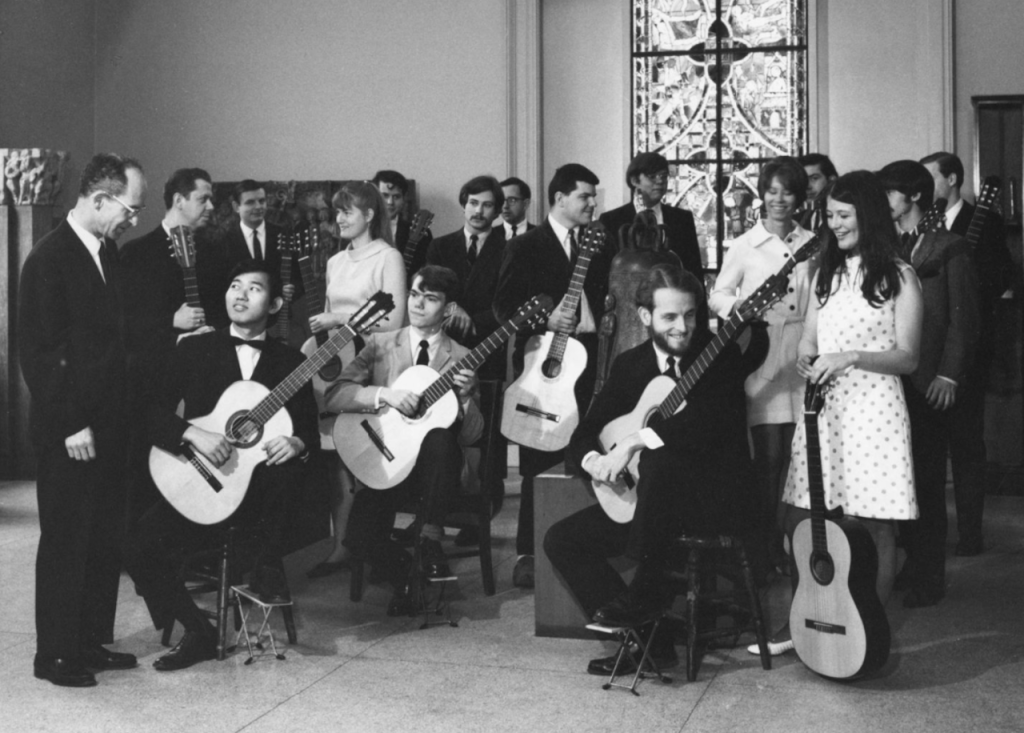A group of people, mixed with some sitting and most standing, hold guitars as they happily converse with each other.