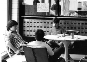 Three students sit at a round table next to old mailboxes in the 1980s. A sign behind them bars students from storing instruments in the security office.