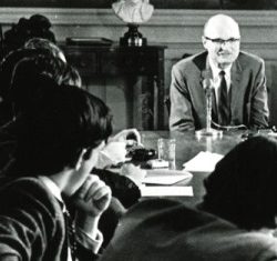 Black and white of students sitting at a wooden table with an older man at the head wearing a suit and tie.