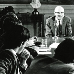 Black and white of students sitting at a wooden table with an older man at the head wearing a suit and tie.