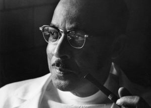 A black man in white coat and oval glasses stares off to the left holding a pipe in one hand.