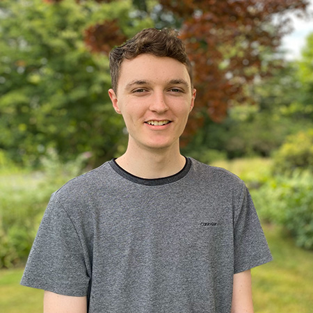 Headshot of a young man wearing a grey shirt with trees, bushes, and grass in the background.