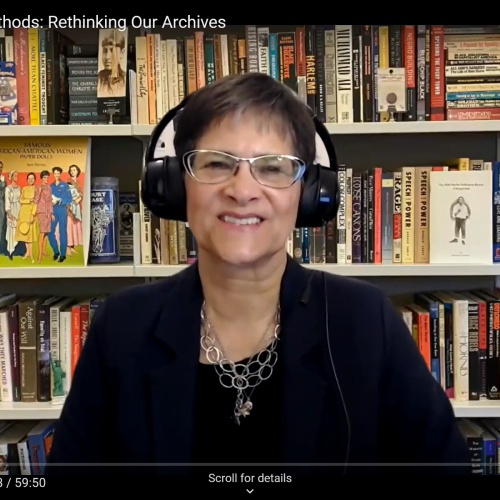 Screenshot of a fair skinned woman on a Zoom call wearing headphones and with a bookcase as her background.