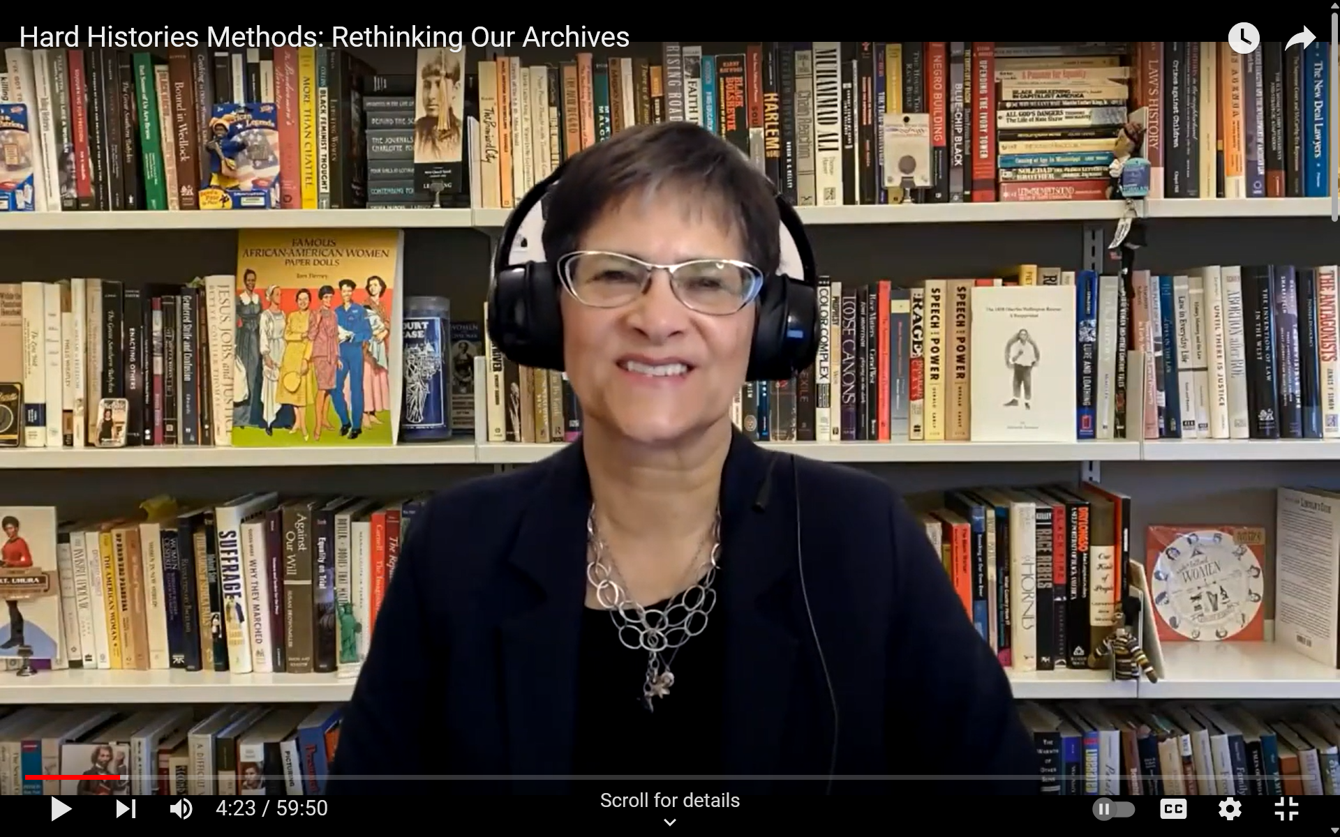 Screenshot of a fair skinned woman on a Zoom call wearing headphones and with a bookcase as her background.