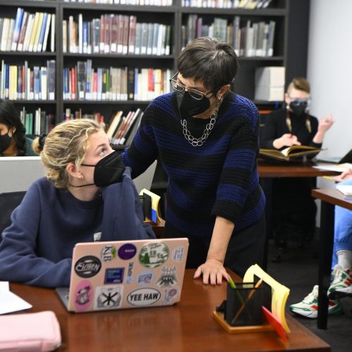 A woman wearing a mask talks to a student sitting in at a table with her laptop open. Both engage in conversation.