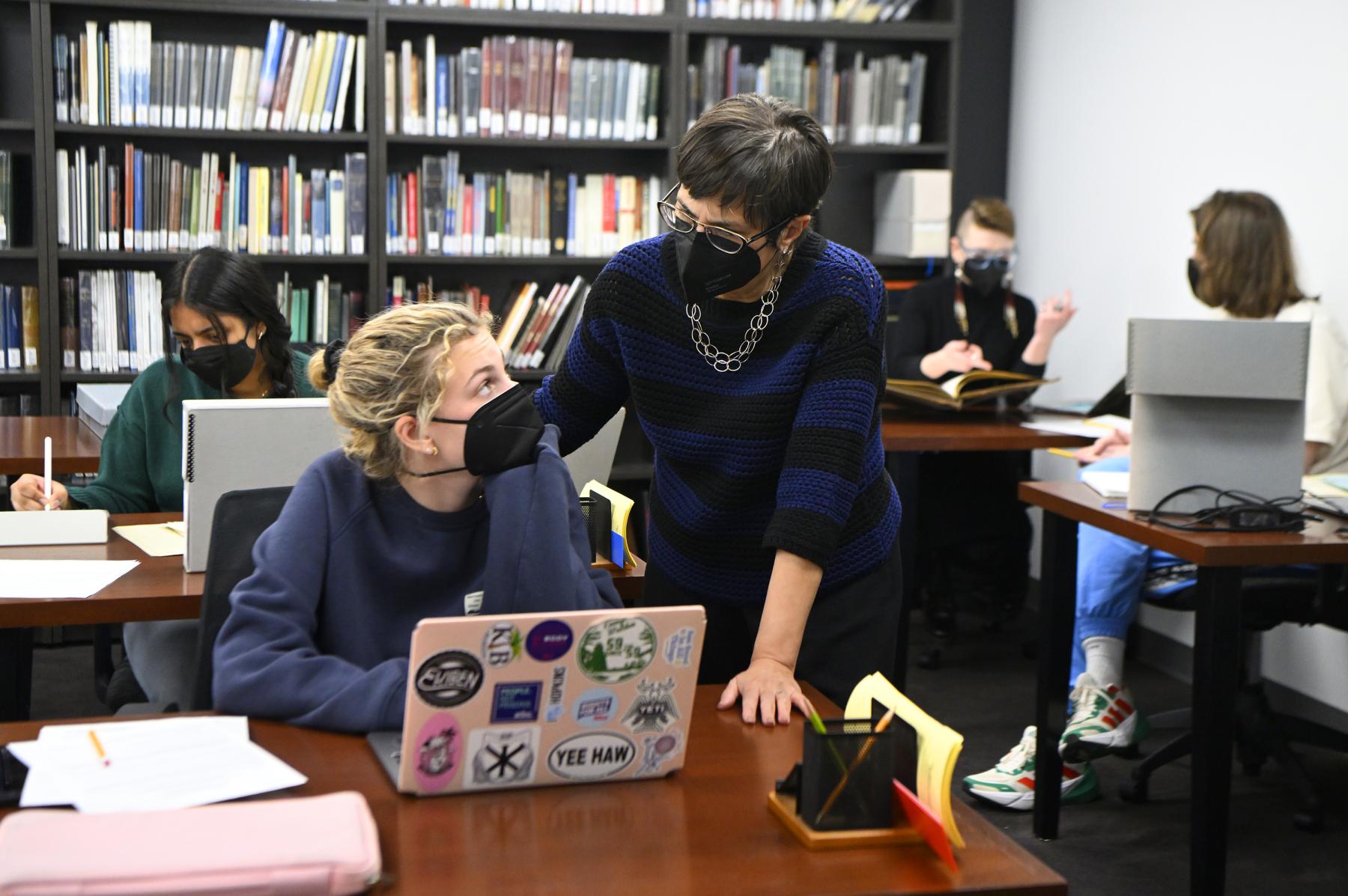 A woman wearing a mask talks to a student sitting in at a table with her laptop open. Both engage in conversation.