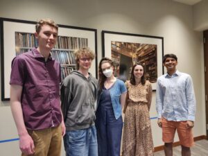 Five college students stand together in front of large framed photos of books on shelves. They smile at the camera and wear semi-formal outfits. 