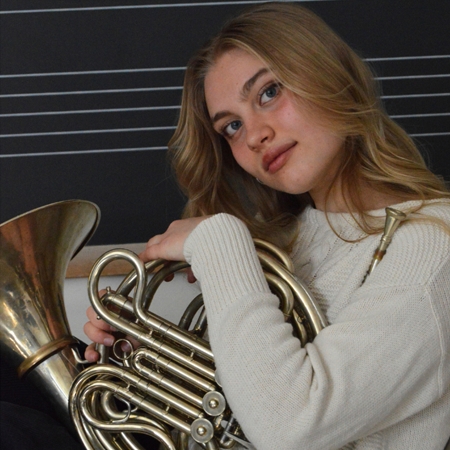 A headshot of a young blonde woman wearing an off-white sweater and holding a French horn close.