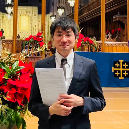 A young man stands in front of a church pulpit, the space decorated with poinsettias. He wears glasses, a suit with a tie, and papers in his hands.