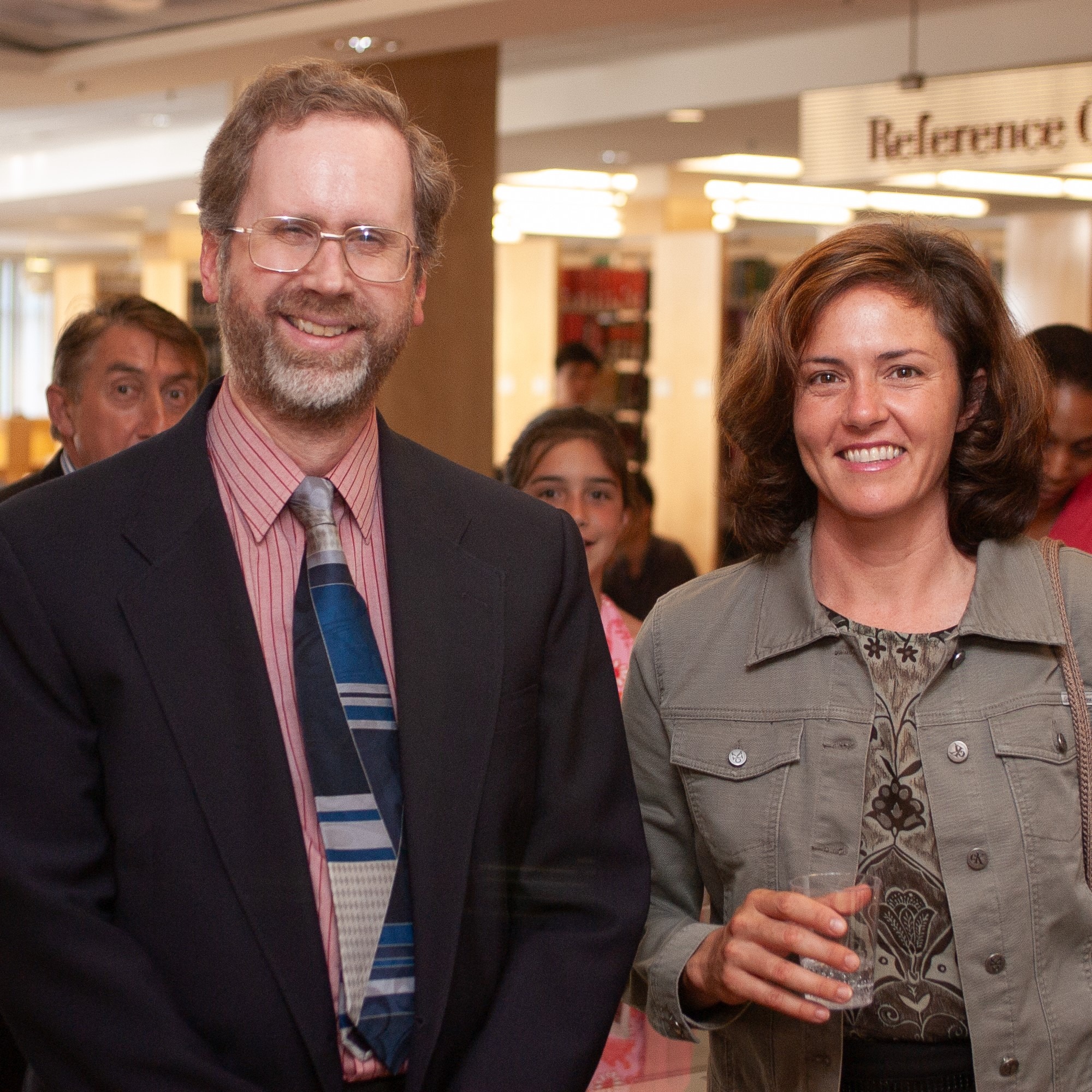 A man and woman stand next to each other and smile at the camera. The man wears a dark suit jacket, striped white and red button up short, and a dark blue patterned tie. The woman wears a jacket and patterned blouse and holds a glass in her hand. They stand in front of a sign that hangs from the ceiling that reads Reference Collection with book stacks beyond the sign. Several other people mill about behind them.