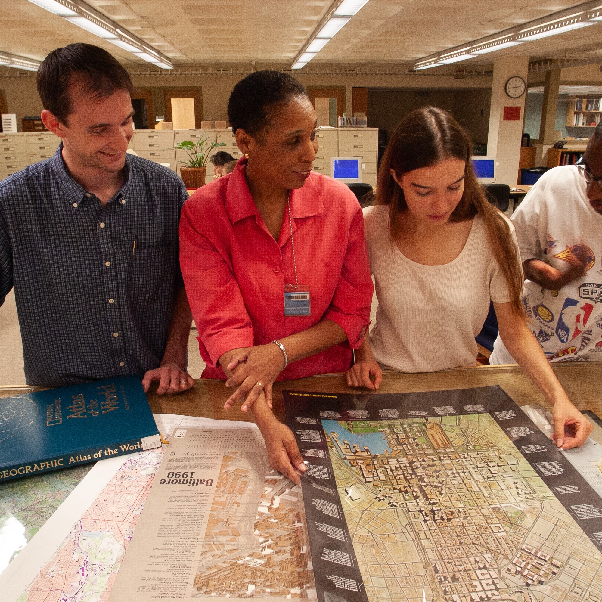 A woman with a short fro shows several maps laid out on top of a low wooden table to three other people standing next to her.