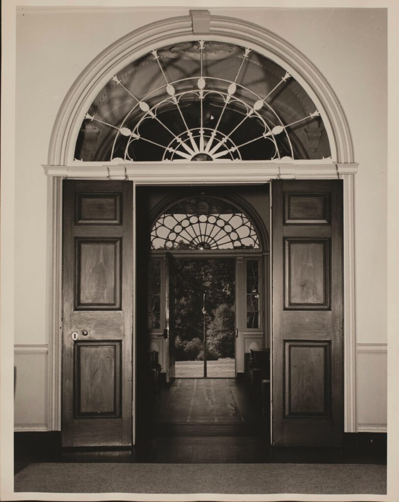 Sepia-toned photo of two open wooden doorways with large, half-circle transoms decorated with sun motifs. The doors lead out onto a lawn and lush landscape.