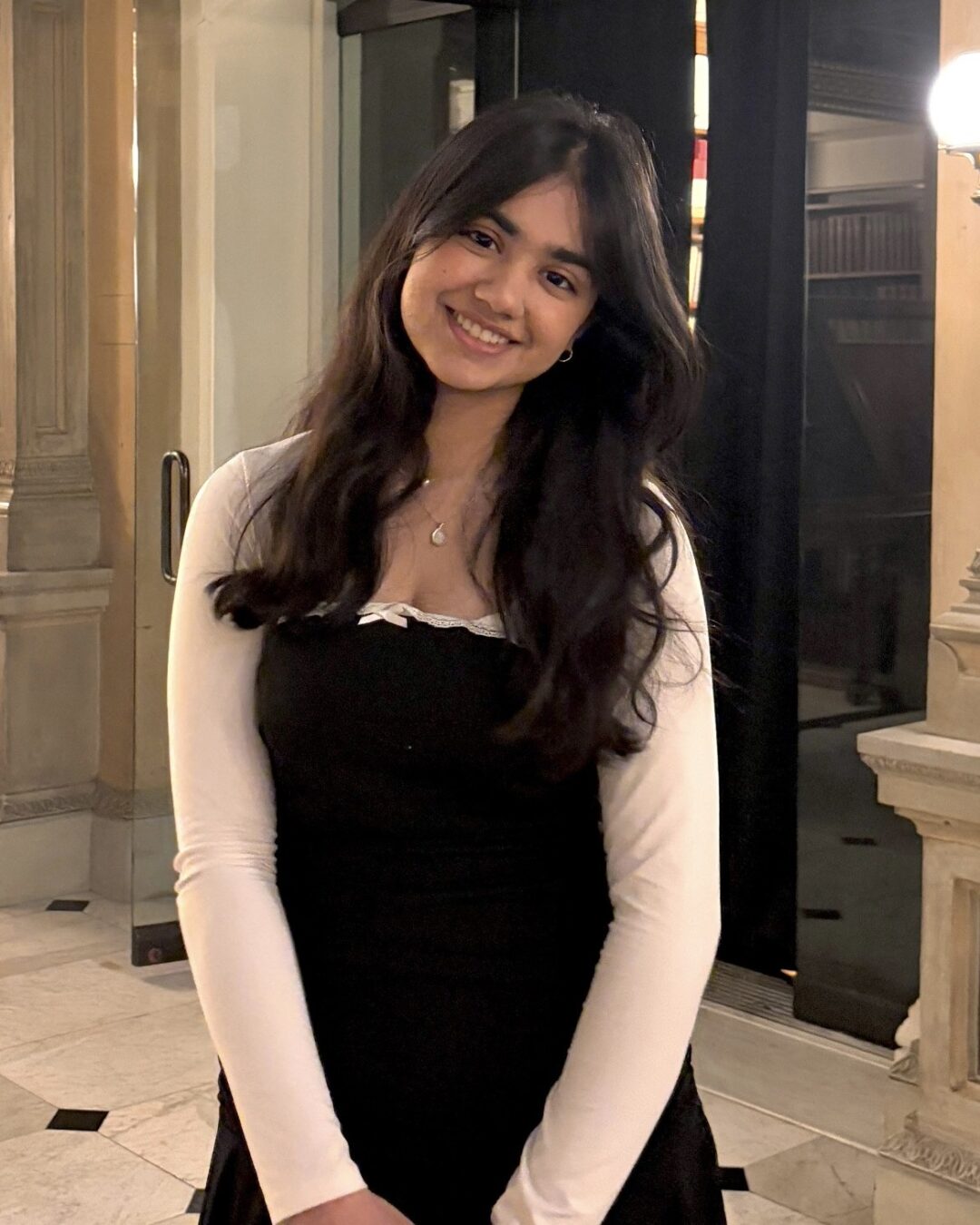Myra Saeed headshot Three-quarter photo of a young woman with long dark hair wearing a black dress with white long sleeves. She smiles at the camera and stands in a upscale lobby.