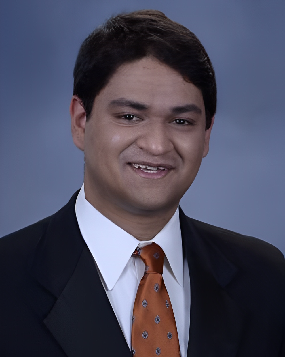 Shaan Bhandarkar headshot Headshot of a young man smiling and wearing a suit jacket with a red tie.